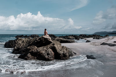 Rear view of woman sitting on rocky shore against sky