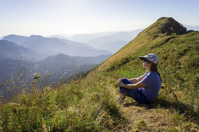 Young woman in cap sitting on mountain against sky
