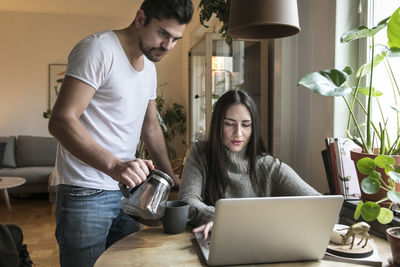 Woman using laptop while man pouring coffee in mug on table