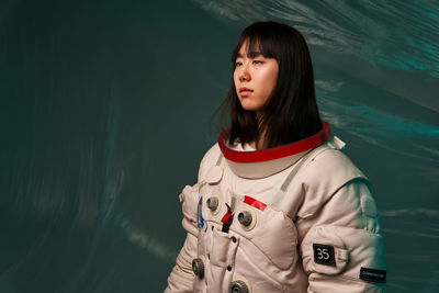 Serious young asian woman in spacesuit with dark hair looking away while standing against green background