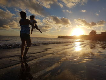 Rear view of woman playing with daughter at beach against sky during sunset