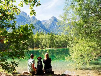 Rear view of people with dog sitting by lake against trees