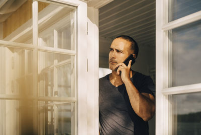 Thoughtful man talking on smart phone while standing at window