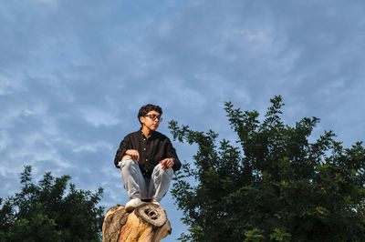 Young transgender man with glasses posing on a log outdoors.