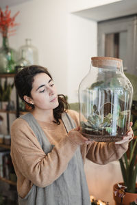 Side view of female in casual clothes holding big transparent glass terrarium closed with cork lid and looking at small green succulents inside while standing against blurred interior of modern flower shop