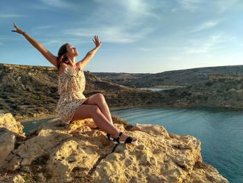 Young woman sitting on rock against sky