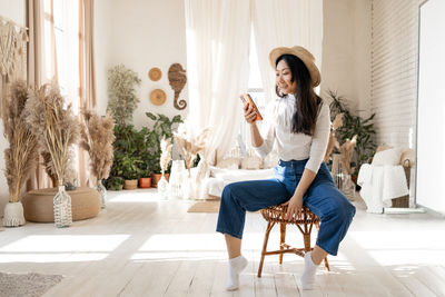 Stylish asian woman in jeans sits on a chair using smartphone over a bed and house plants