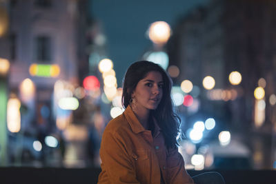 Portrait of young woman sitting in illuminated city at night