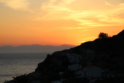 Silhouette buildings by sea against orange sky