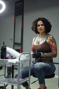 Serious adult woman in casual clothes with tattoos sitting in light tattoo salon while looking at camera