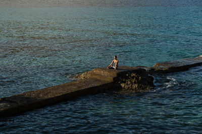 Peaceful female sitting on stone in padmasana and meditating with mudra gesture near sea during sundown