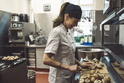 Side view of smiling female chef cutting mushrooms at kitchen counter