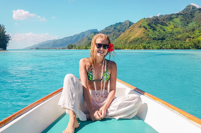 Portrait of woman wearing sunglasses while sitting in boat on sea against mountains and sky