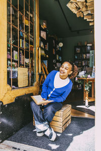 Smiling woman holding book while sitting on crate near door at bookstore
