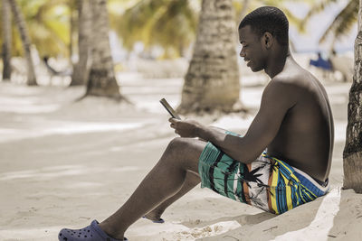 Shirtless black male traveler smiling and browsing social media on cell phone while sitting