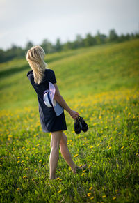 Rear view of young woman with hat holding sandals on grassy field