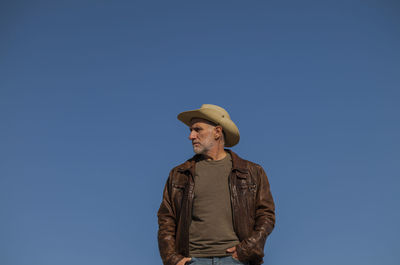 Portrait of adult man with brown leather jacket and cowboy hat against blue clear sky