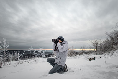 Man photographing while kneeling on field against sky during winter