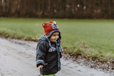 Cute boy wearing in warm clothing standing on dirt road by field