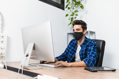 Unrecognizable young stylish male entrepreneur in face mask working on desktop computer in modern workspace