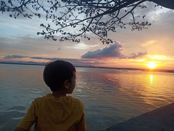 Rear view of boy looking at sea against sky during sunset