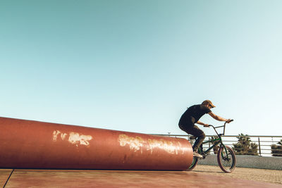Man riding bicycle by red metallic pipe against sky