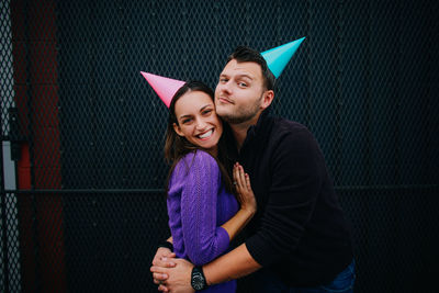 Portrait of man embracing woman while wearing party hats against metal grate