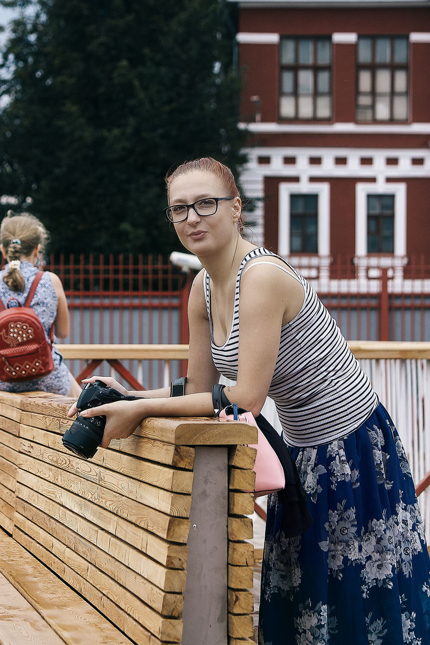 glasses, sitting, architecture, eyeglasses, real people, built structure, lifestyles, building exterior, leisure activity, young adult, people, casual clothing, young women, day, women, focus on foreground, front view, three quarter length, outdoors, hairstyle