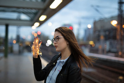 Young woman holding illuminated string lights while standing at railroad station platform at dusk