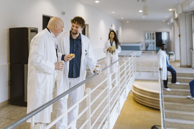 Happy male physicians talking while standing by railing during coffee break in hospital corridor