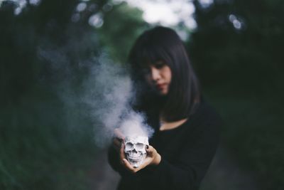 Woman holding human skull emitting smoke against trees