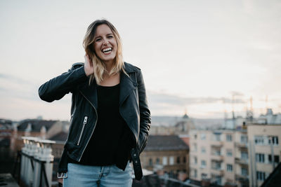 Portrait of smiling young woman standing against cityscape during sunset