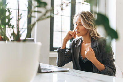 Young blonde woman with long hair in stylish suit working at laptop in office, talking on video call