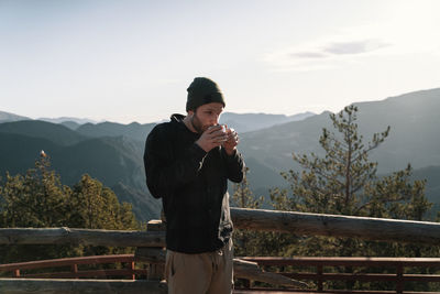 Man standing on railing against mountains drinking coffee