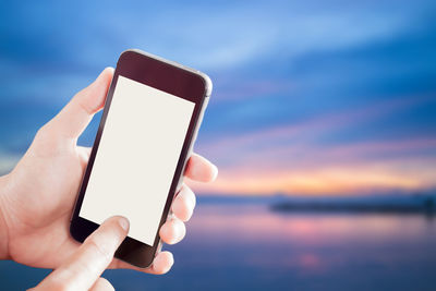 Close-up of hand holding smart phone against sky during sunset