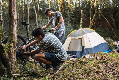 Side view of male cyclist fixing mechanism of bicycle with friend against hiker setting up tent in forest