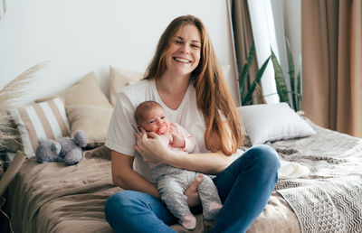 A young laughing mother holds a newborn baby in her arms while sitting on the bed.