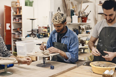 Young man using work tool by friend molding clay while standing at table in art class