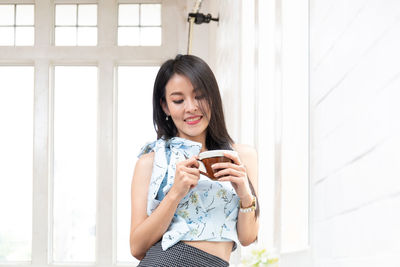 Full length of young woman holding coffee cup by window