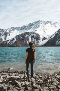 Rear view full length of woman standing at lakeshore against snowcapped mountain