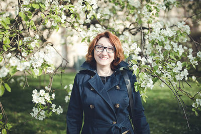 Confident middle aged armenian woman in a blue trench coat and glasses under the blooming tree