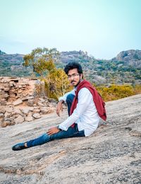 Side view portrait of young man sitting on rock against sky