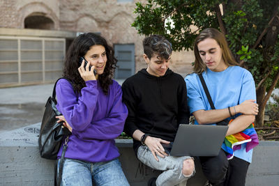 Young diverse people with lgbt rainbow flag using laptop and mobile outdoors.