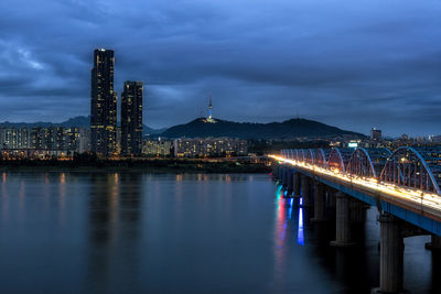 Illuminated bridge over river by buildings against sky at dusk