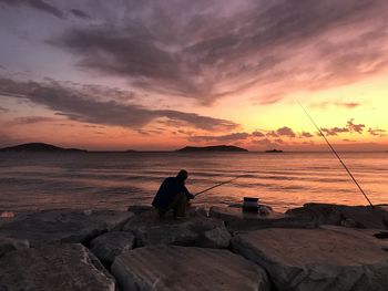 Man fishing on rock by sea against sky during sunset