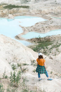 Back view of young woman traveler in cap  standing on shore blue water of clay quarry and sand 