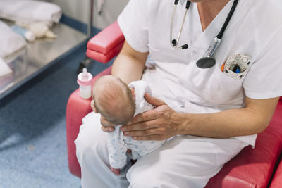 Doctors sitting on armchairs and feeding newborns from bottles holding heads in room with equipment in hospital