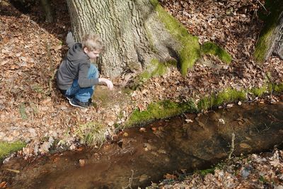 Boy sitting by tree trunk at stream