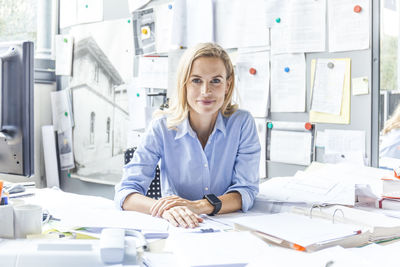 Portrait of confident woman sitting at desk in office surrounded by paperwork