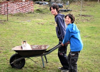 Boy holding wheelbarrow with cat while standing by brother on field